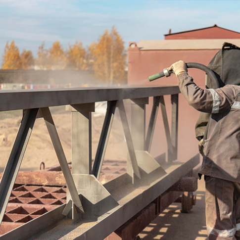 Industrial-Sandblasting-in-Progress-for-Structural-Steel-Component فني يرتدي معدات الحماية يقوم بضرب الرمل على عارضة فولاذية لتحضير سطحها للطلاء – يبرز خدمات مؤسسة ASC في تجهيز الأسطح الصناعية