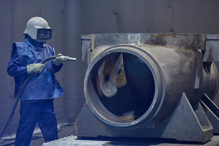 Sandblasting-Specialist-Preparing-Heavy-Metal Workpiece عامل صناعي يستخدم أداة ضرب الرمل لمعالجة قطعة معدنية كبيرة – يبرز احترافية مؤسسة ASC في تجهيز الأسطح